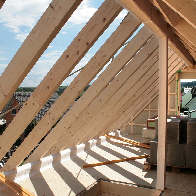 Charpente en bois d'un toit en construction, avec vue sur des maisons et le ciel. Des blocs de béton sont empilés.