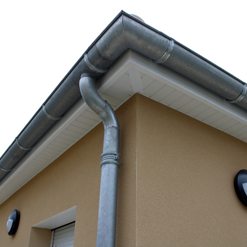 Coin de bâtiment avec gouttière métallique et descente d'eau grise sur une façade beige claire, sous un soffite blanc.
