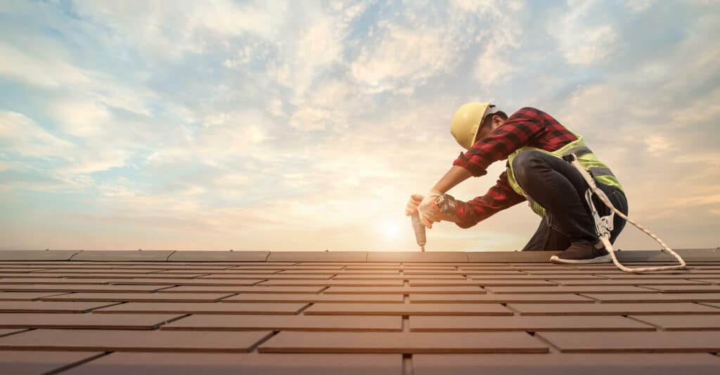 Ouvrier avec casque et harnais de sécurité installant des tuiles marron sur un toit au coucher du soleil.