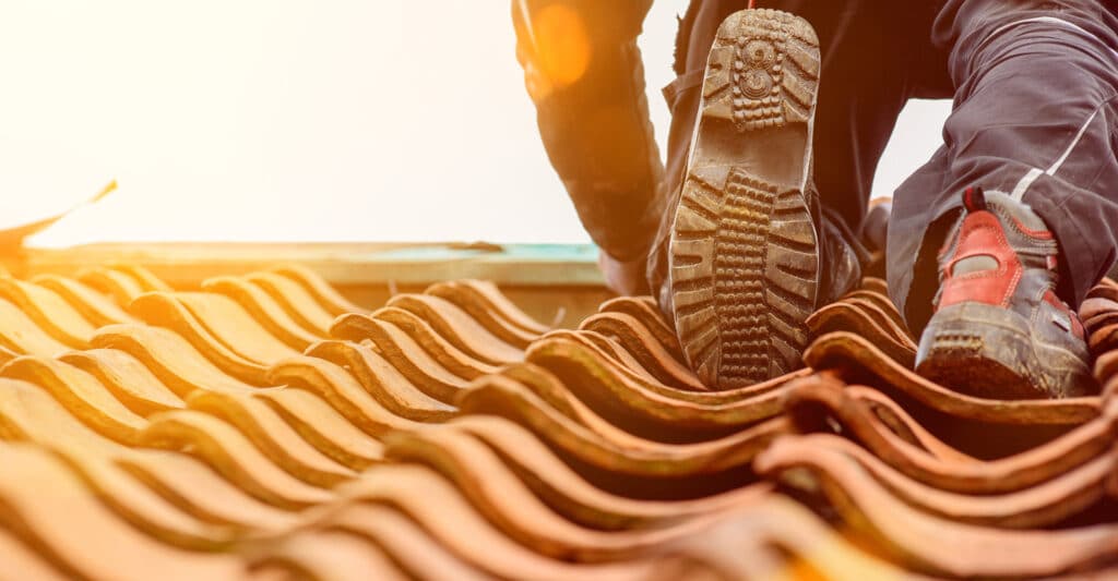 Ouvrier en chaussures de sécurité sur un toit de tuiles en terre cuite, en plein soleil. Vue sur la semelle d'une botte et le bout d'une chaussure.