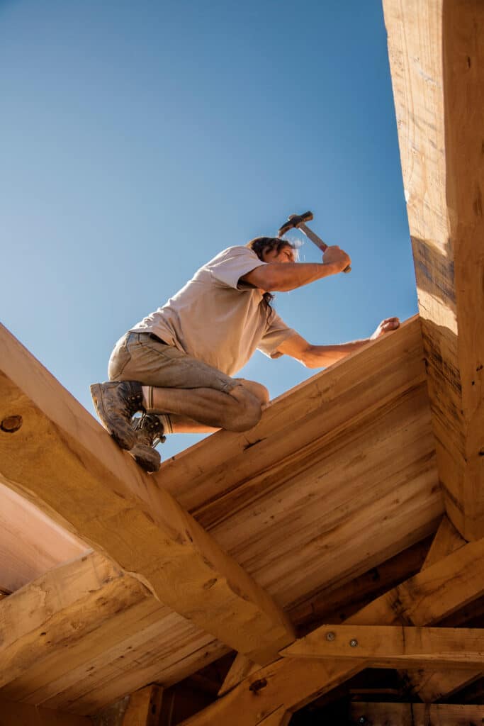 Charpentier agenouillé sur une structure en bois, le ciel bleu en arrière-plan. Il lève un marteau pour frapper une poutre.