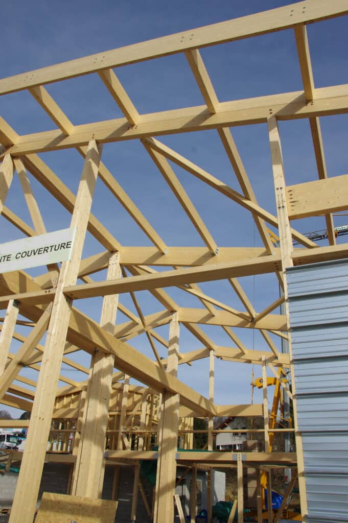 Grande charpente en bois d'un bâtiment en construction sous ciel bleu. Détail des poutres, boulons et panneau "NTE COUVERTURE".
