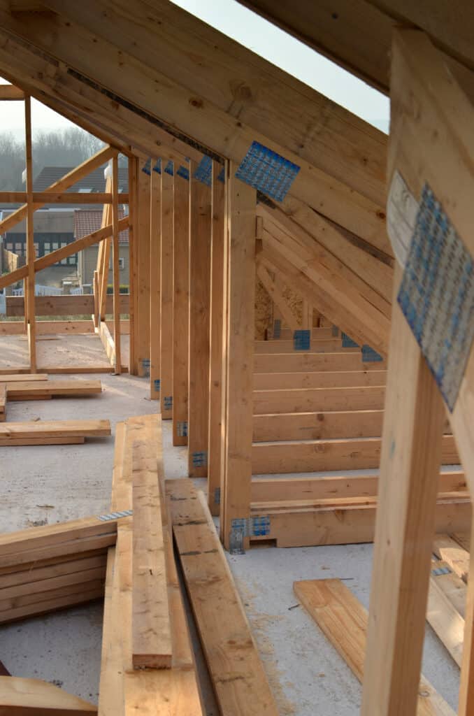 Intérieur de la charpente en bois d'une maison en construction, avec poutres et montants sur une dalle béton. Matériaux au sol et maisons en arrière-plan.