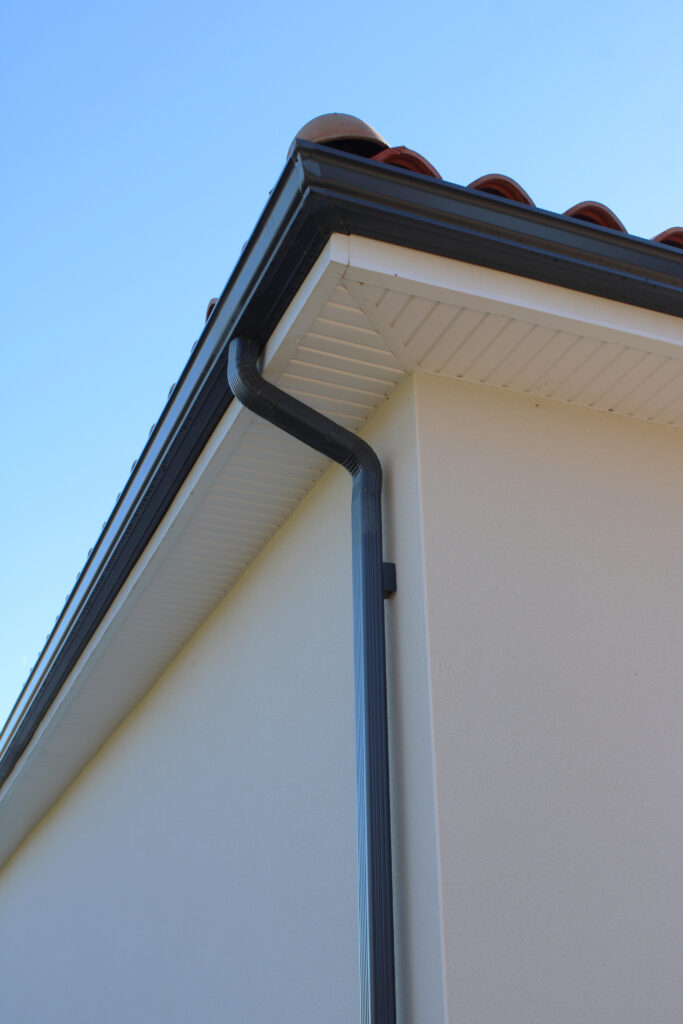 Gouttière sombre et descente pluviale sur un coin de maison moderne beige avec tuiles rouges et ciel bleu clair.