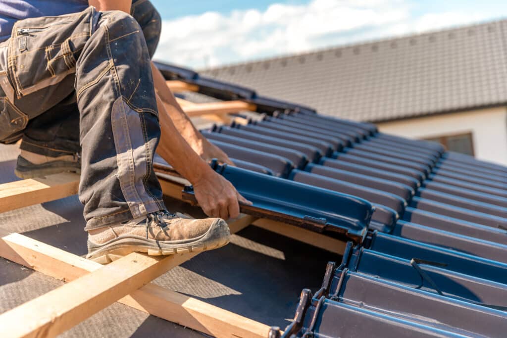Roofer en bottes de travail posant des tuiles foncées sur une nouvelle toiture. Gros plan sur les mains et les tuiles sous un ciel ensoleillé.
