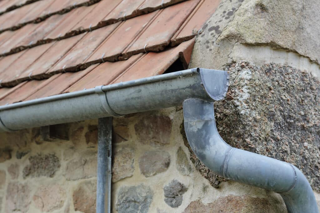 Gouttière en zinc grise sous des tuiles de toit en terre cuite usées sur un mur de pierre rustique d'une ancienne maison.